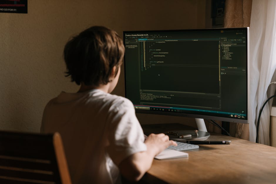 A teenager focused on coding software on a desktop monitor in a home office setting.